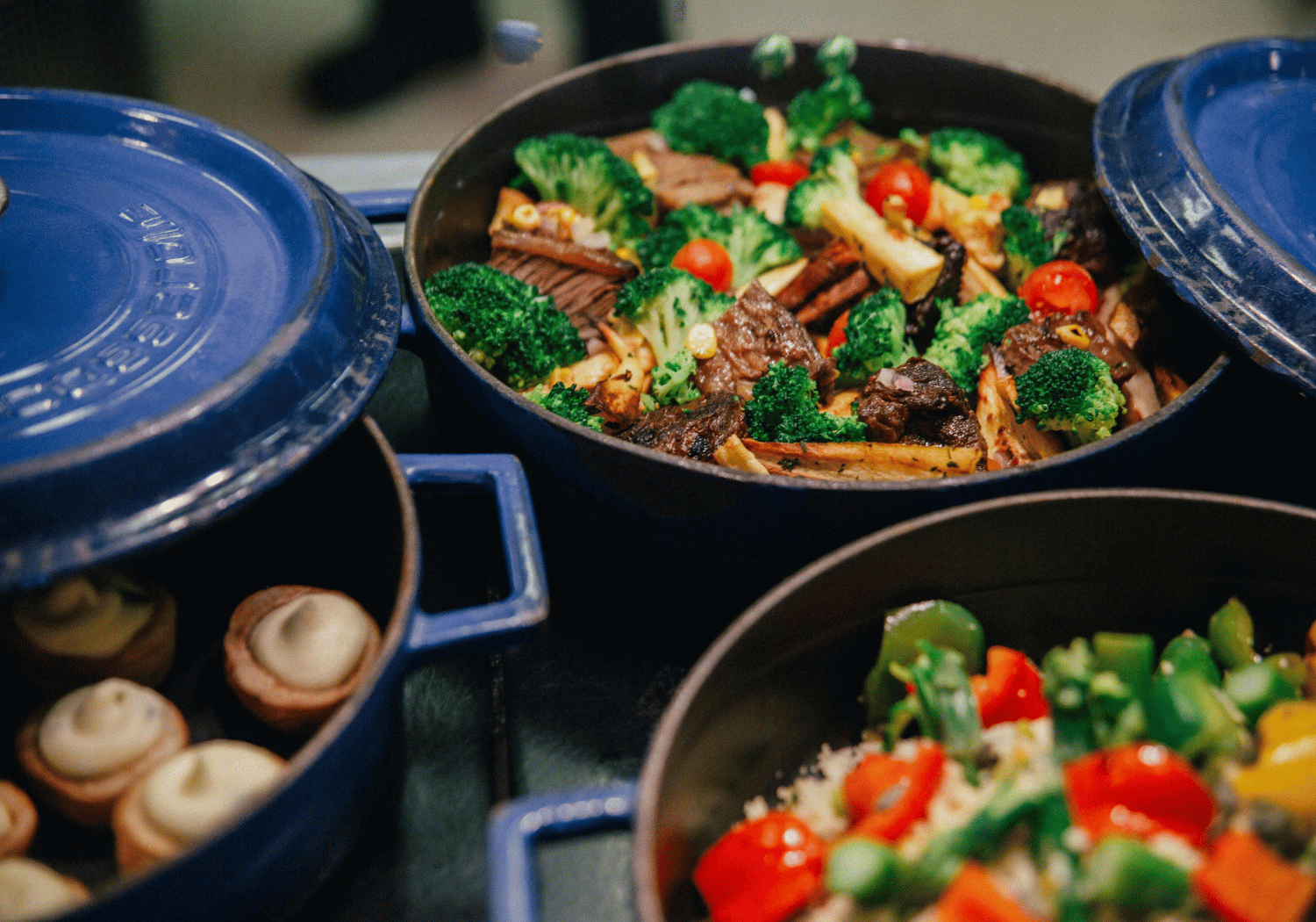 Blue pot filled with beef and broccoli stir-fry with cherry tomatoes and potatoes, framed by other blue pots on a table